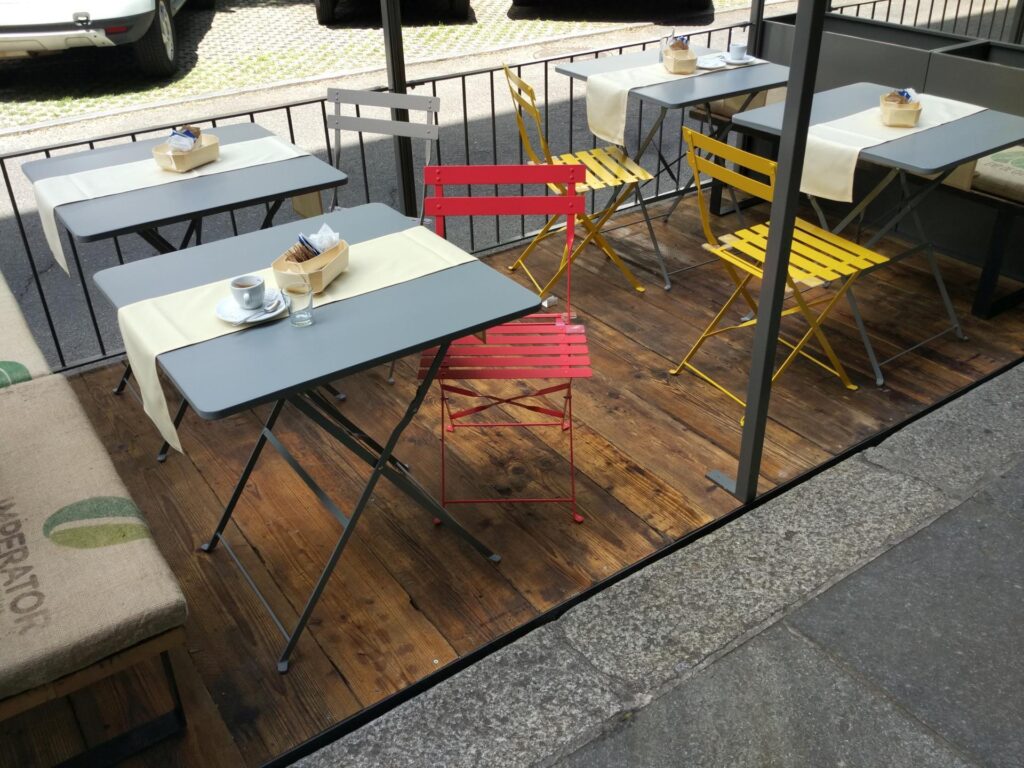 Studio di Progettazione La Manifattura. Arredamento di un dehors esterno, con pavimento in tavole di legno, tavolini quadrati smaltati di colore grigio chiaro e seggioline pieghevoli in ferro smaltato di colore rosso, giallo e grigio. Oltre i tavolini, verso strada, c'è una ringhiera in ferro di colore grigio scuro.
