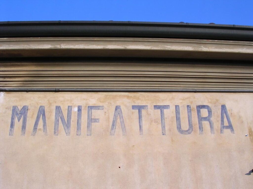 Studio di Progettazione La Manifattura. Un cielo azzurro e sotto un muro color ocra con la scritta in stampatello MANIFATTURA