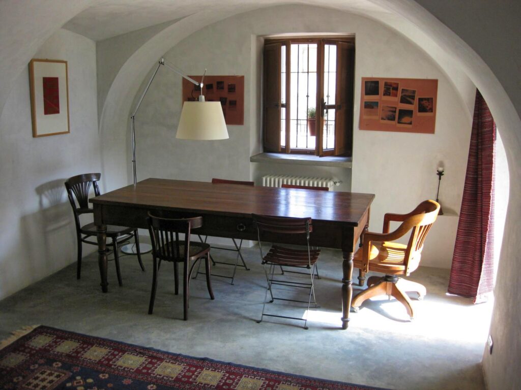 Studio di Progettazione La Manifattura. Arredamento di un soggiorno, con sedie tutte diverse e posizionate intorno a un tavolo rettamgolere in legno. I soffitti sono bianchi e a volta, oltre il tavolo c'è una finestra in legno con gli scuri interni. in primo piano, sul pavimento in cemento c'è un tappeto con motivi geometrici.