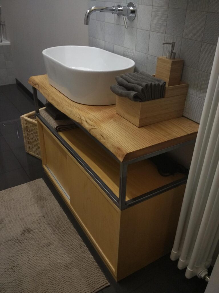 Studio di Progettazione La Manifattura. Arredamento cottituito dal mobile in legno di un bagno su cui si vede un lavandino in appoggio, di colore bianco e con i lati arrotondati. Sulla parere piastrelle di colore grigio con piccolissimi motivi geometrici. Sul pavimento si vede un tappeto di colore beige,